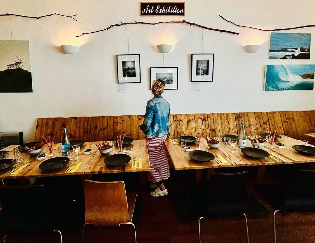 Sala interior com mesas corridas de madeira postas para a refeição e fotografias expostas na parede